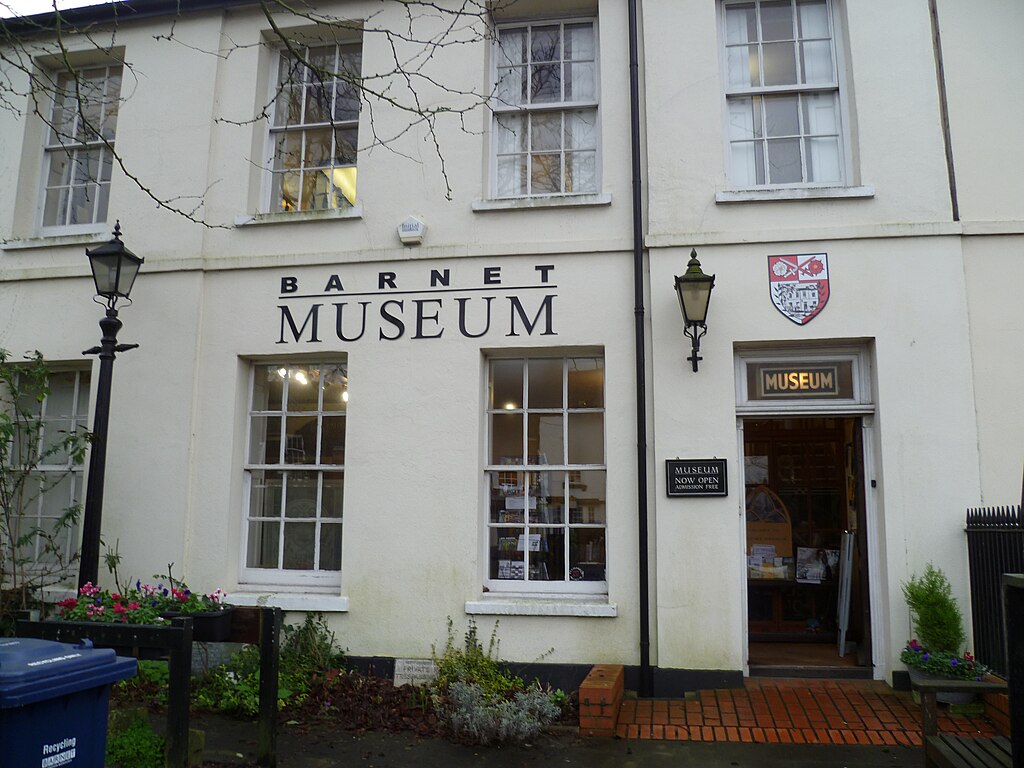 Exterior of Barnet Museum with open door and signage, flanked by lanterns and a coat of arms. | Philafrenzy, CC BY-SA 4.0, via Wikimedia Commons