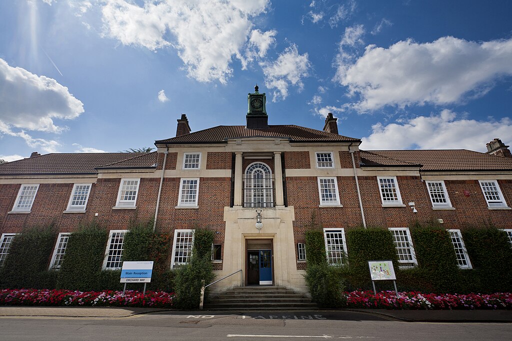 Brick building with a clock tower, blue sky above, and flower bed in front. | SLaMNHSFT, CC BY-SA 3.0, via Wikimedia Commons