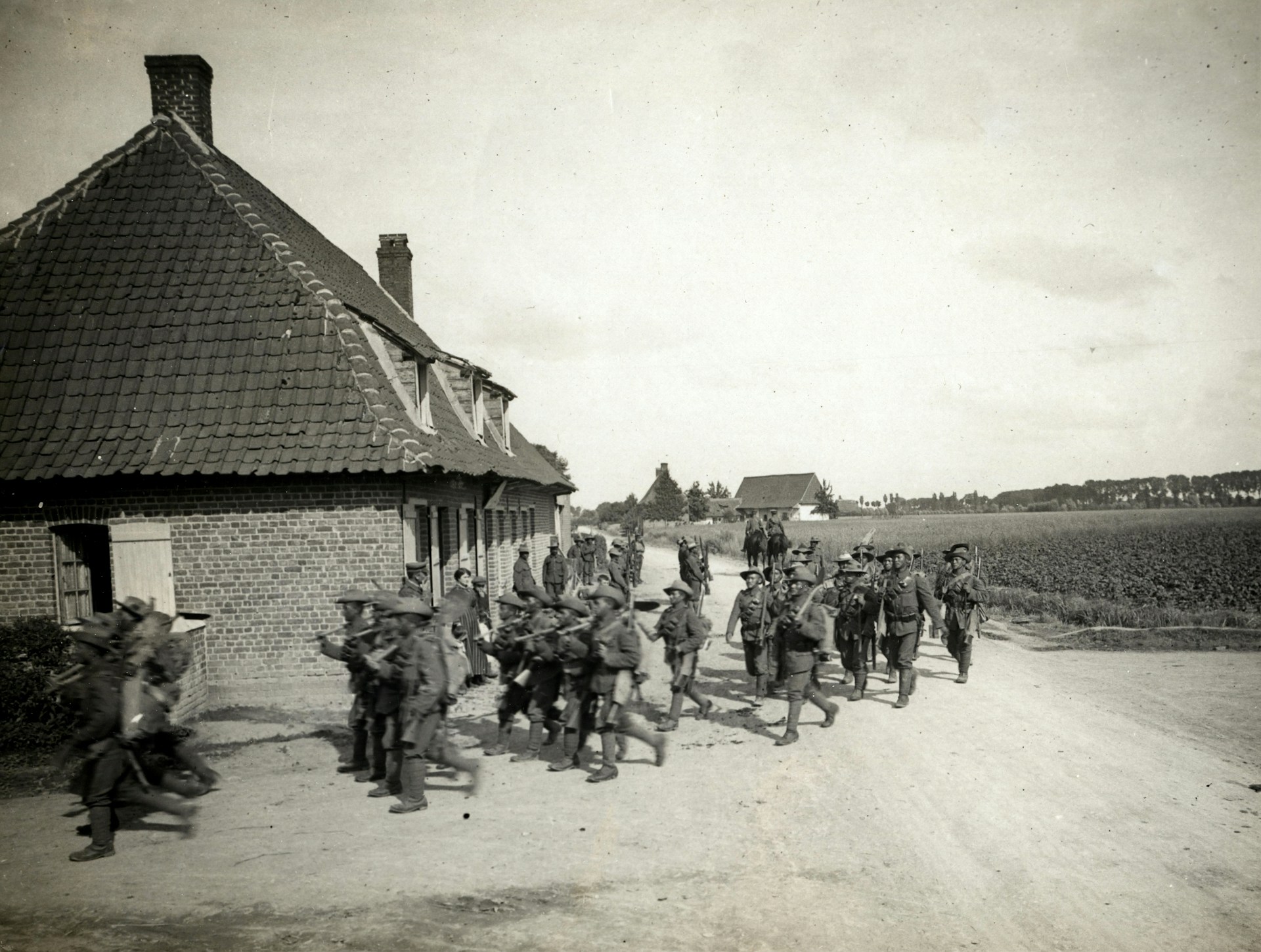 Soldiers marching past a brick house on a dirt road, possibly from an early 20th-century conflict. | Photo by British Library on Unsplash