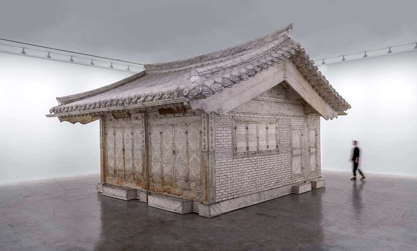 A person walking past a full-scale traditional Korean house sculpture in a gallery.