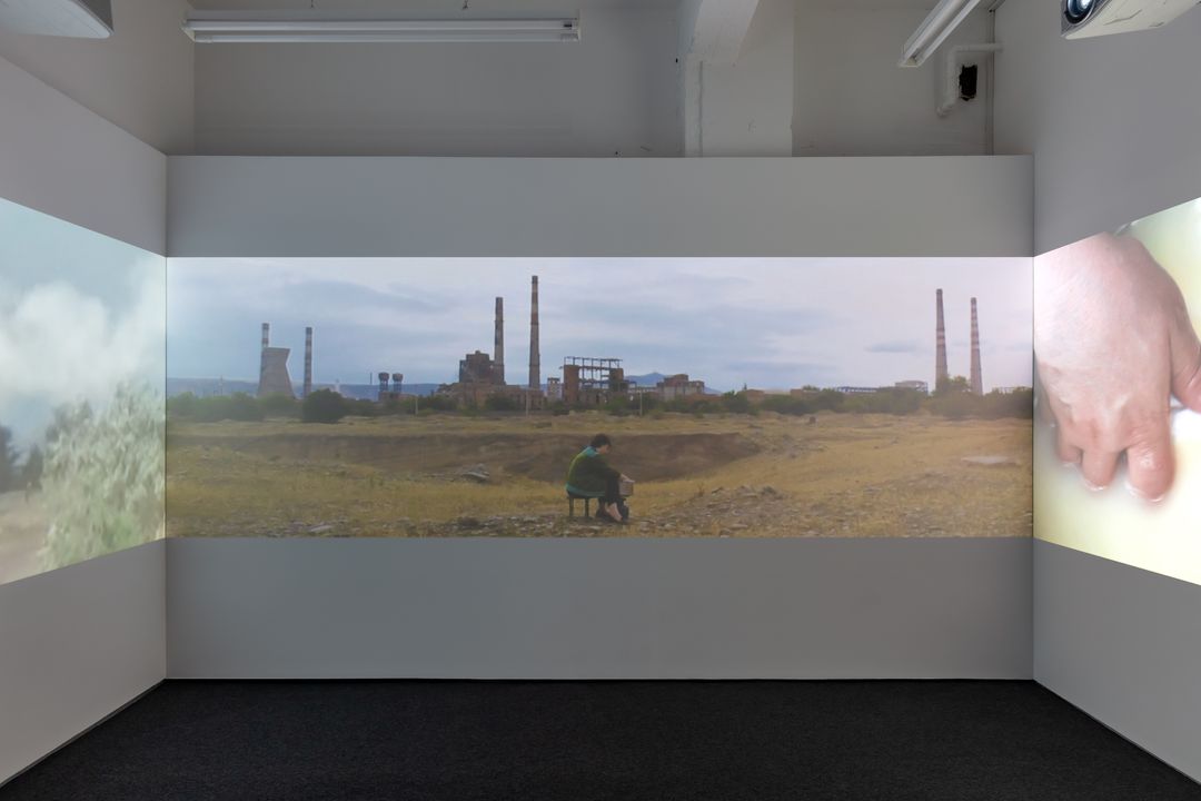Three-panel video installation showing an industrial landscape and a person sitting. | My Mother, Rustavi, 2023