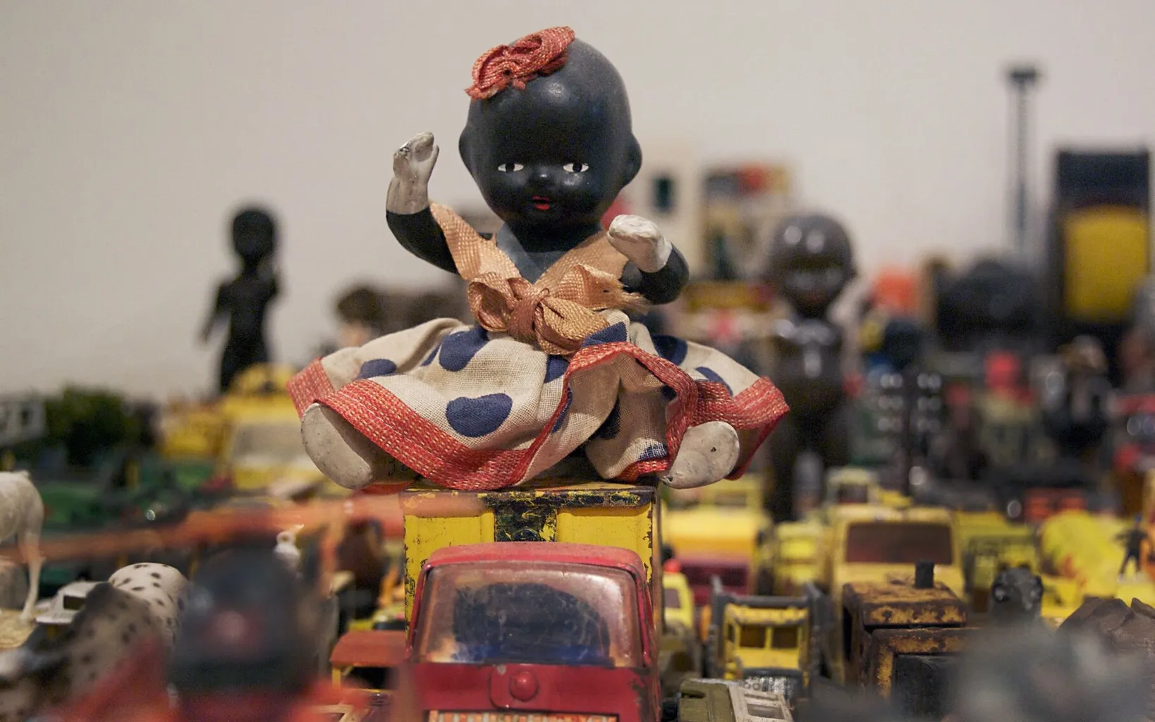 A vintage doll in a polka dot dress sits atop a pile of old toy cars, surrounded by various other miniature vehicles and figures.