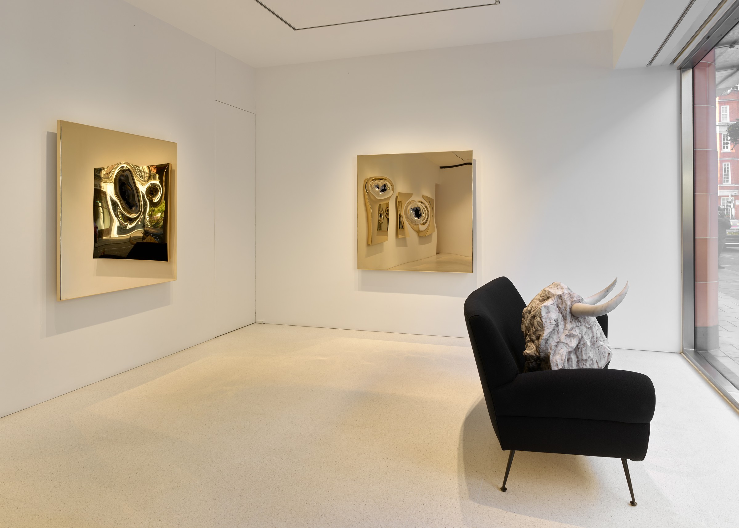 A modern art gallery featuring two reflective paintings on white walls and a black sofa with a sculptural horned element.
