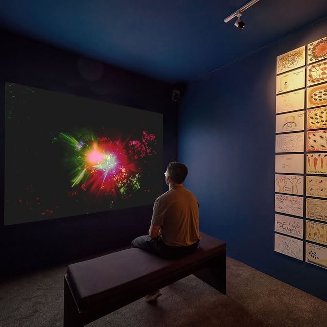 A person sits on a bench, captivated by a vibrant digital art display on a dark wall, surrounded by sketches on another wall.
