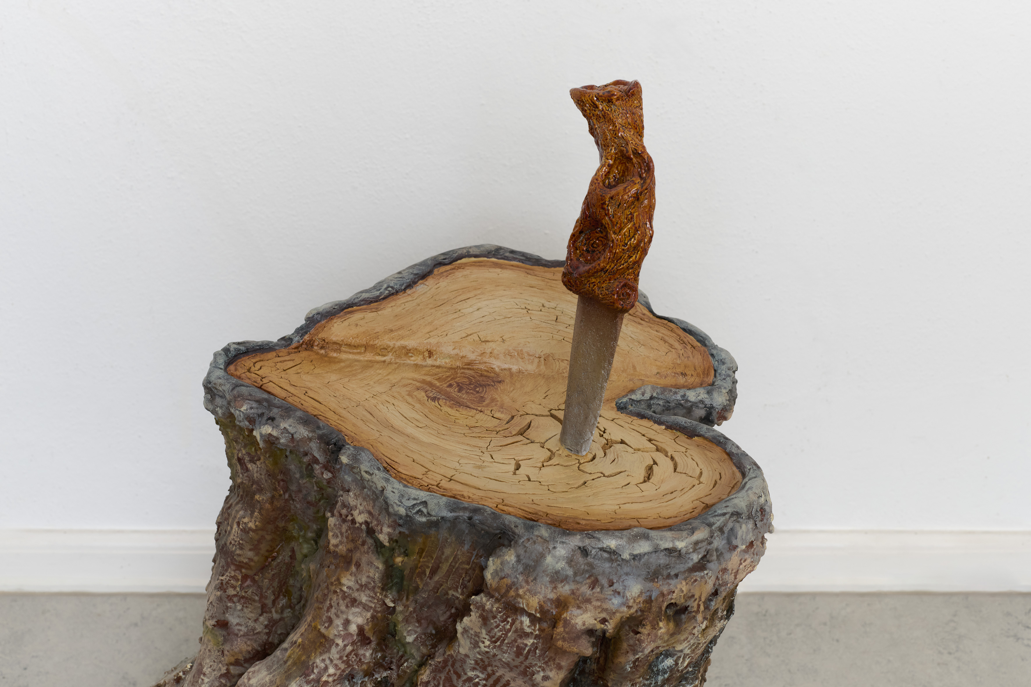A sculpted tree stump with a textured, orange-brown figure emerging from its center, against a minimalist white wall.
