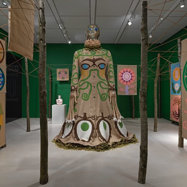 A large, nature-themed sculpture in a vibrant green gallery, surrounded by wooden poles and colorful, abstract art panels.