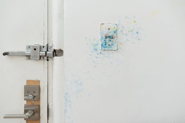 A close-up of a white wall featuring a metal door latch and a faded light switch, with colorful smudges surrounding it.