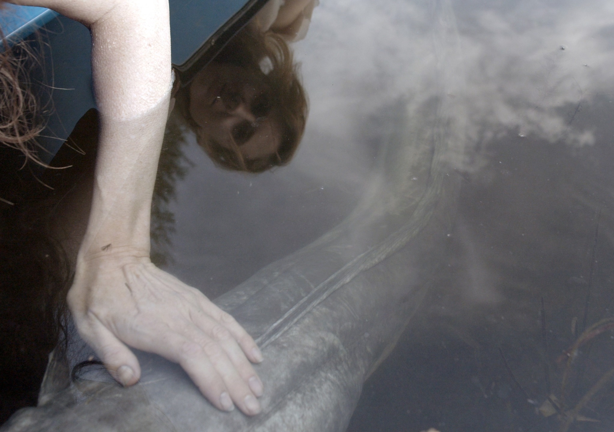 A close-up of a hand touching the surface of dark water, reflecting a woman's face and abstract shapes beneath.
