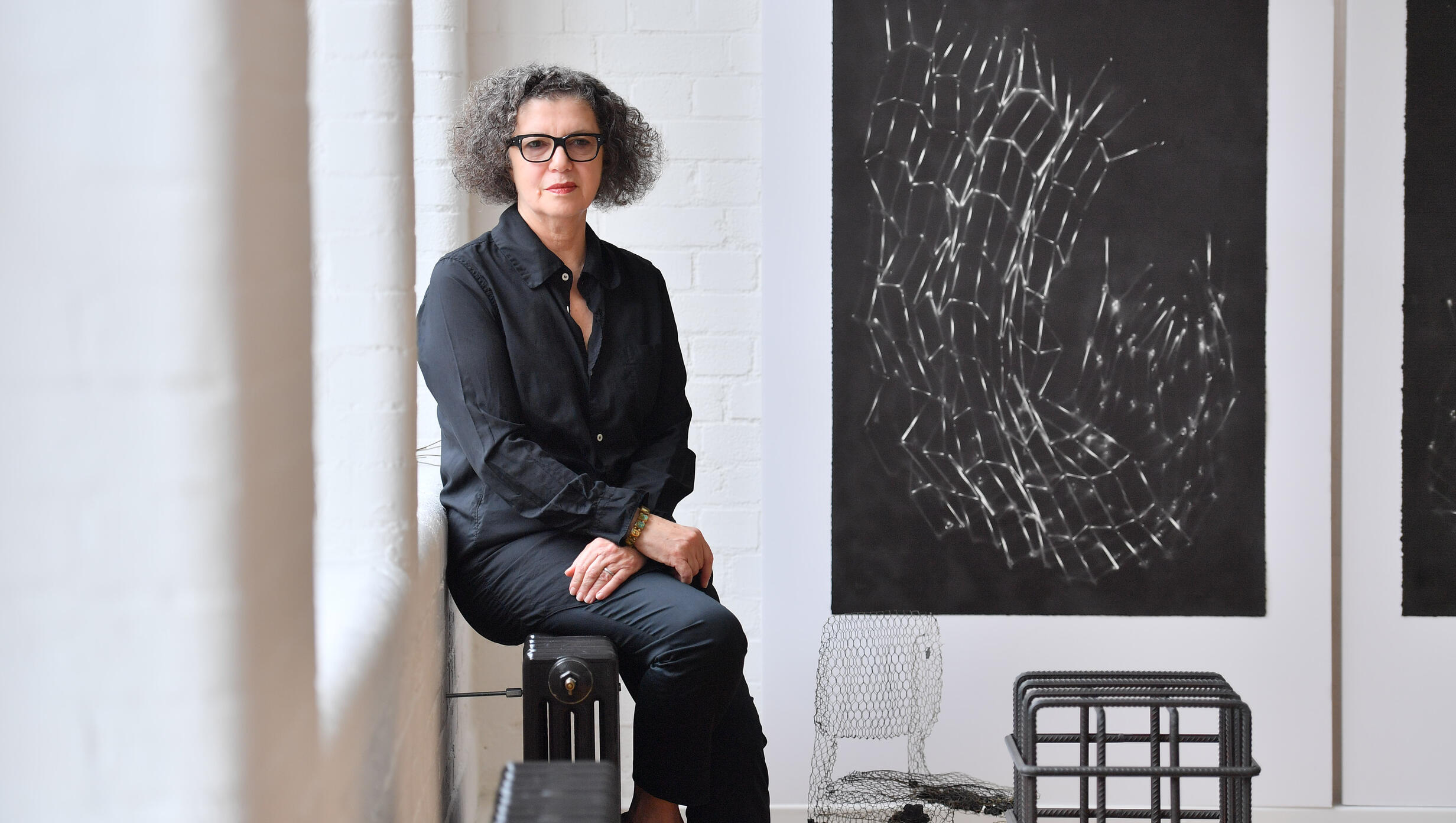 A woman sits on a stool, gazing at a large black and white painting displayed on the wall behind her.