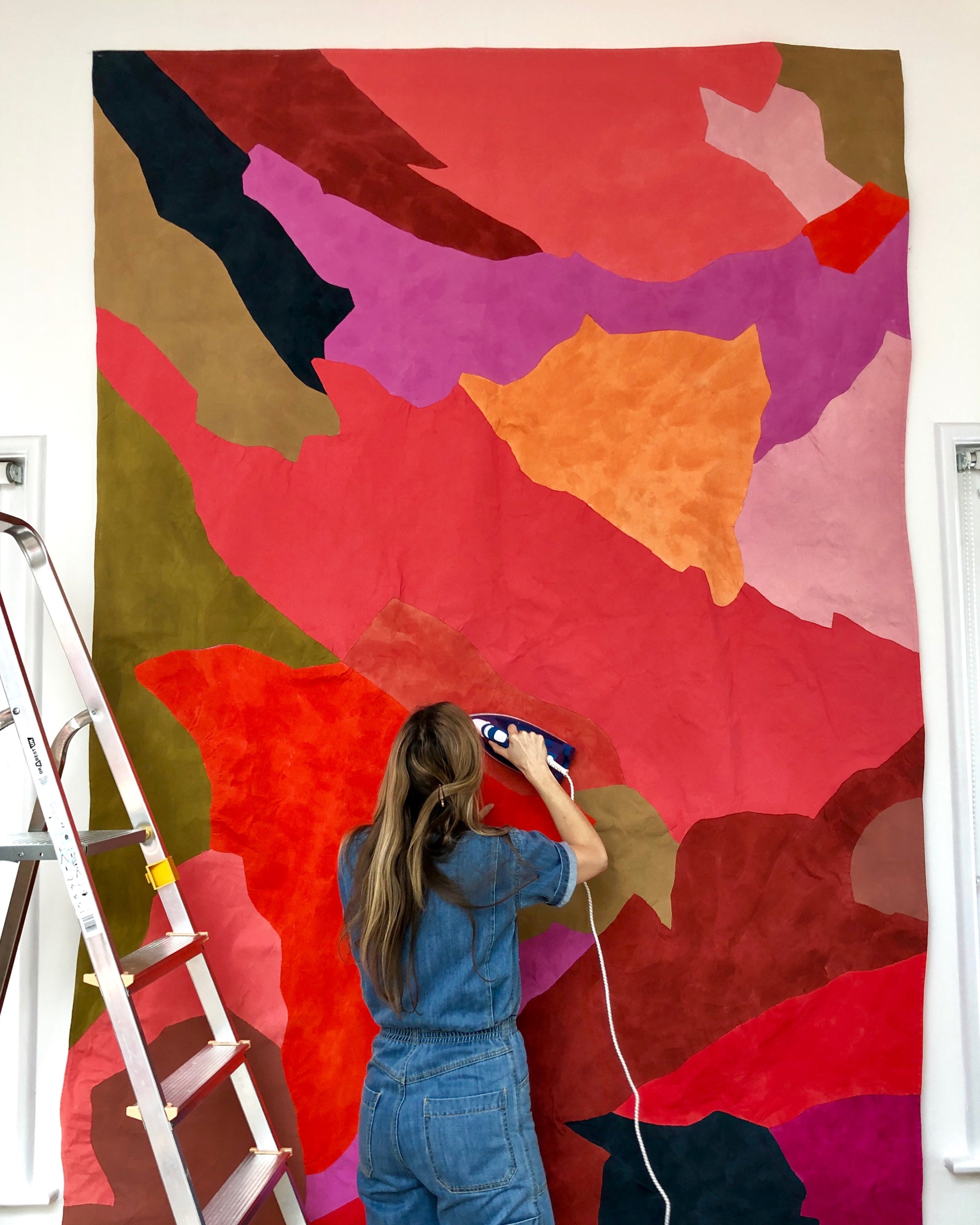 A woman in denim overalls irons a large, colorful fabric mural featuring vibrant abstract shapes in shades of pink, red, and green.