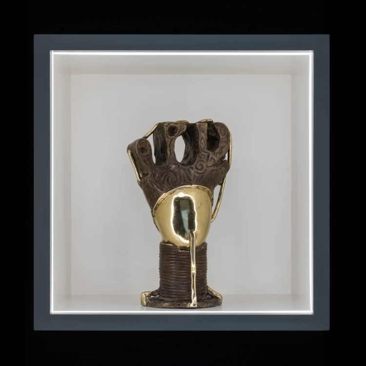A sculptural artwork depicting a raised fist, blending textured bronze and shiny gold elements, displayed in a lighted box.