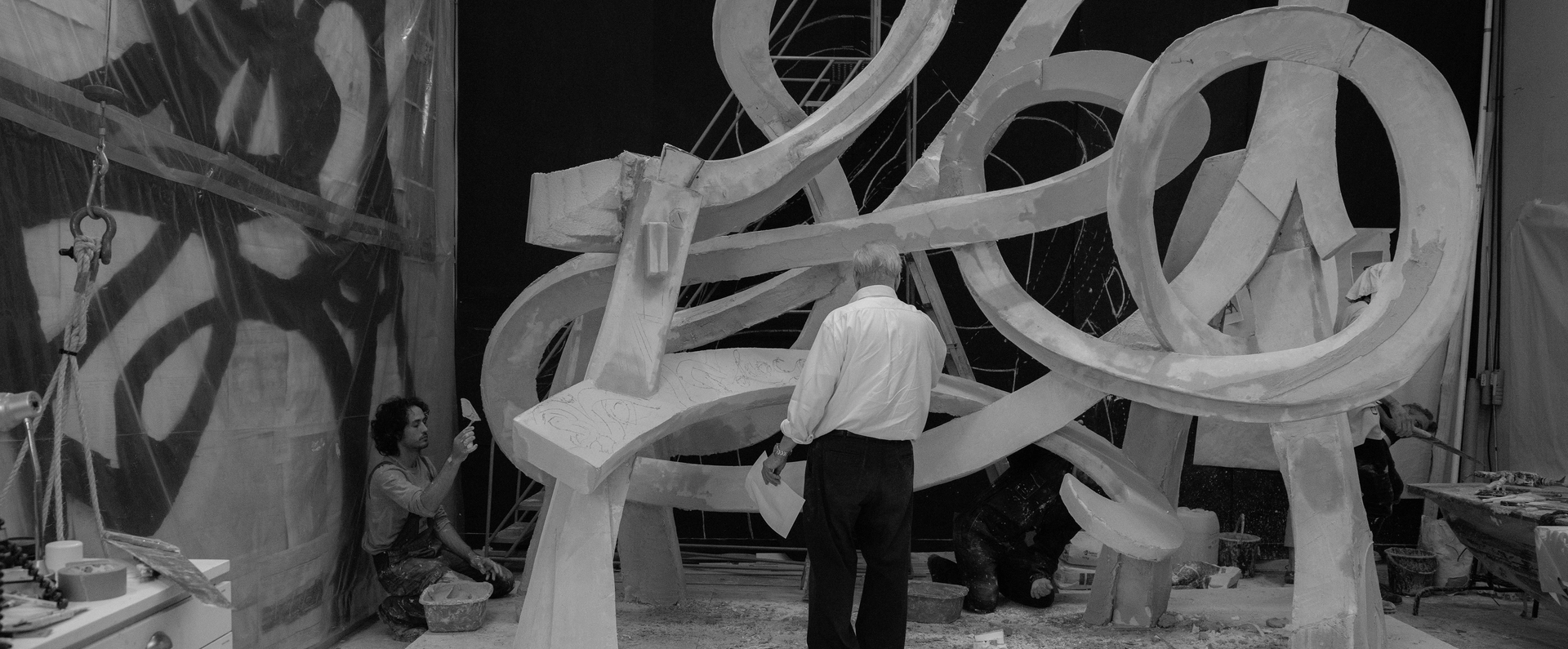An artist examines a large, abstract sculpture in a workshop, with others working diligently on details in the background.