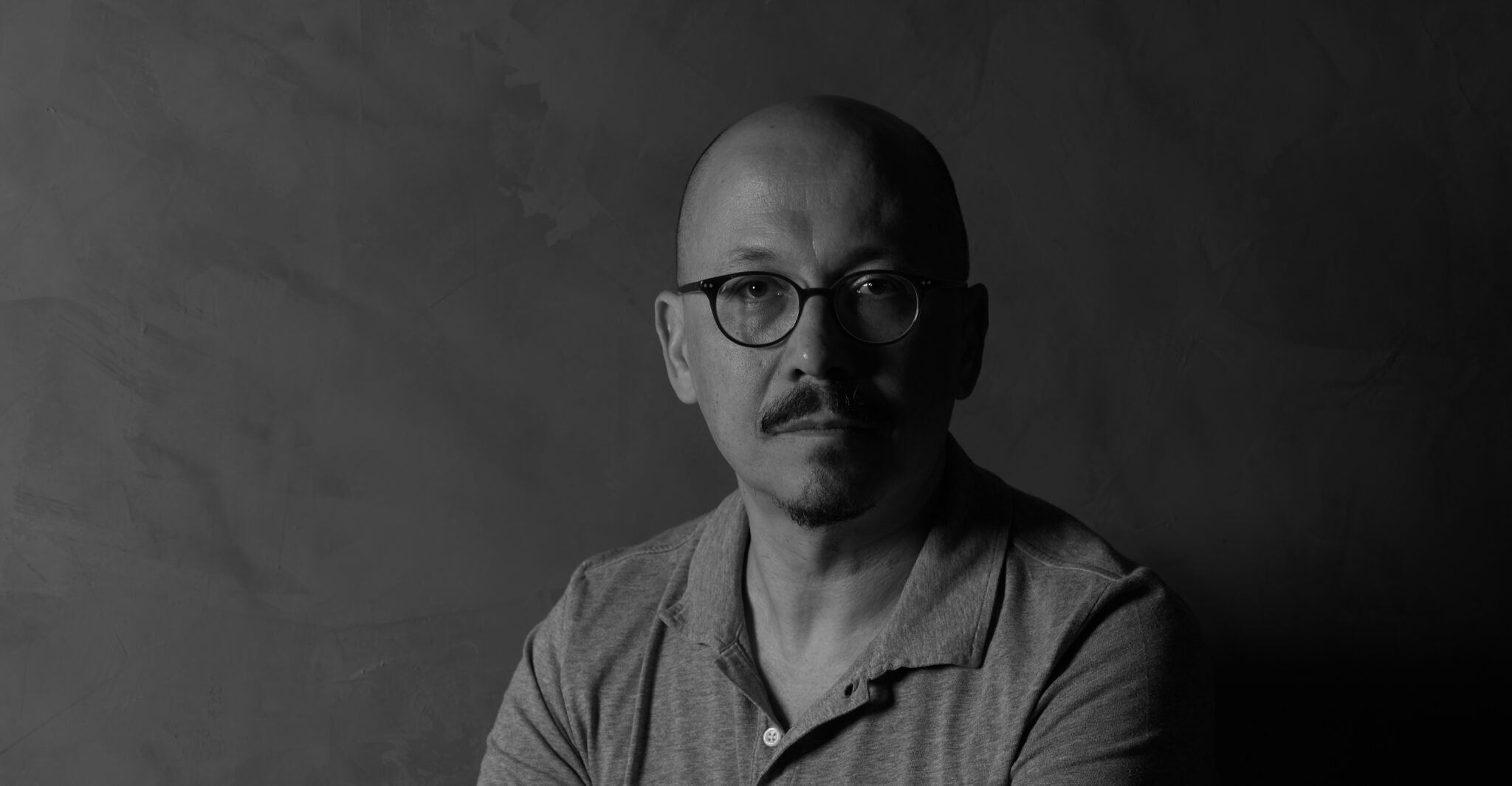 A man with a bald head sits against a dark textured background, wearing a grey polo shirt, creating a somber atmosphere.