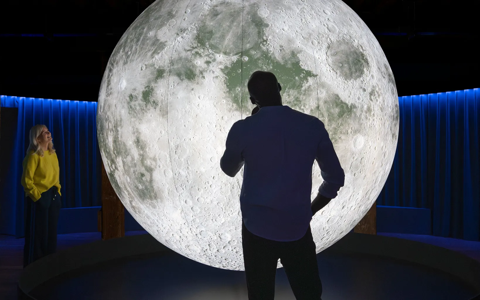 A silhouetted figure stands before a massive, illuminated model of the moon, showcasing its detailed craters and surface features.