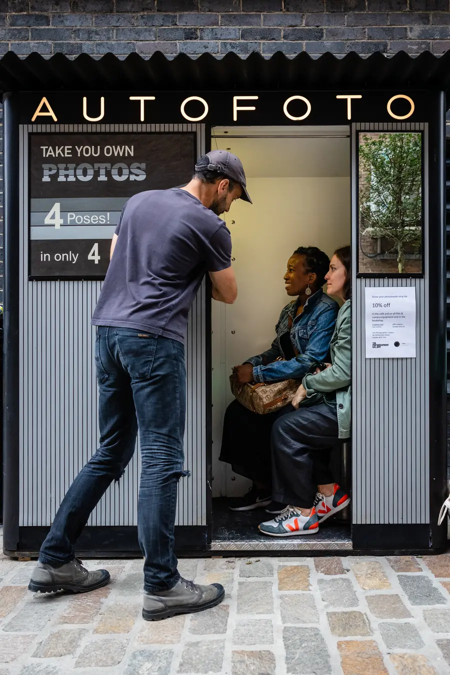 A man assists customers inside a photo booth advertising "AUTOFOTO" with a sign promoting a four-pose photo session for a discount.