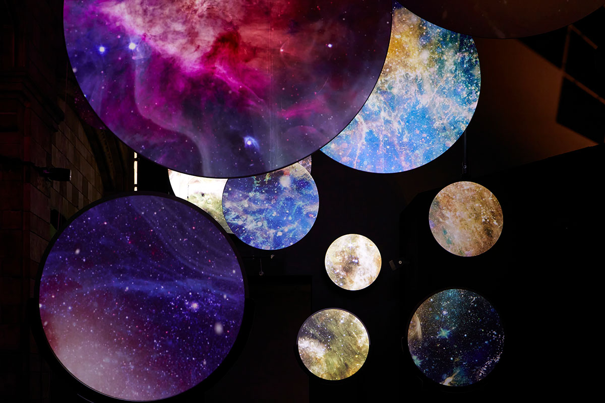 A series of illuminated circular displays feature vibrant, swirling patterns of galaxies and cosmic imagery in a darkened space.