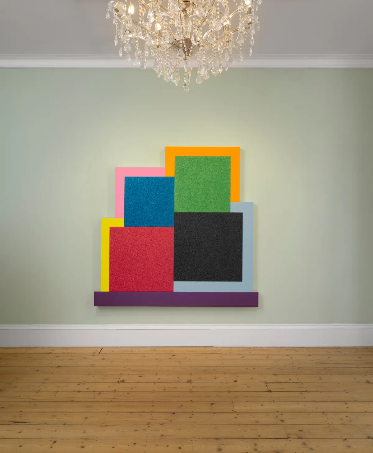 A colorful abstract wall art featuring layered geometric shapes on a light green wall, underneath a sparkling chandelier.
