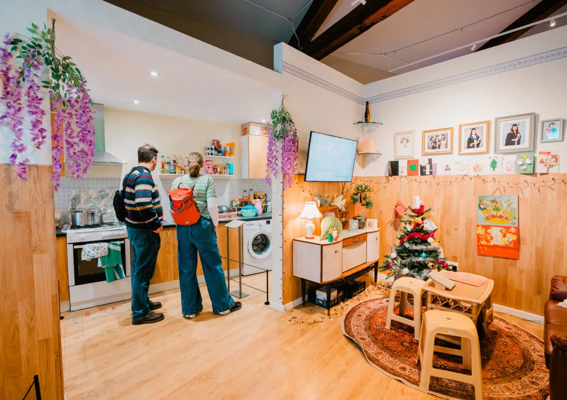 A couple of people standing in a kitchen filled with modern furniture, including a table and chairs. The walls are decorated, and the room has an inviting atmosphere with various art pieces and cabinetry. The flooring is visible beneath them, contributing to the overall home interior design.