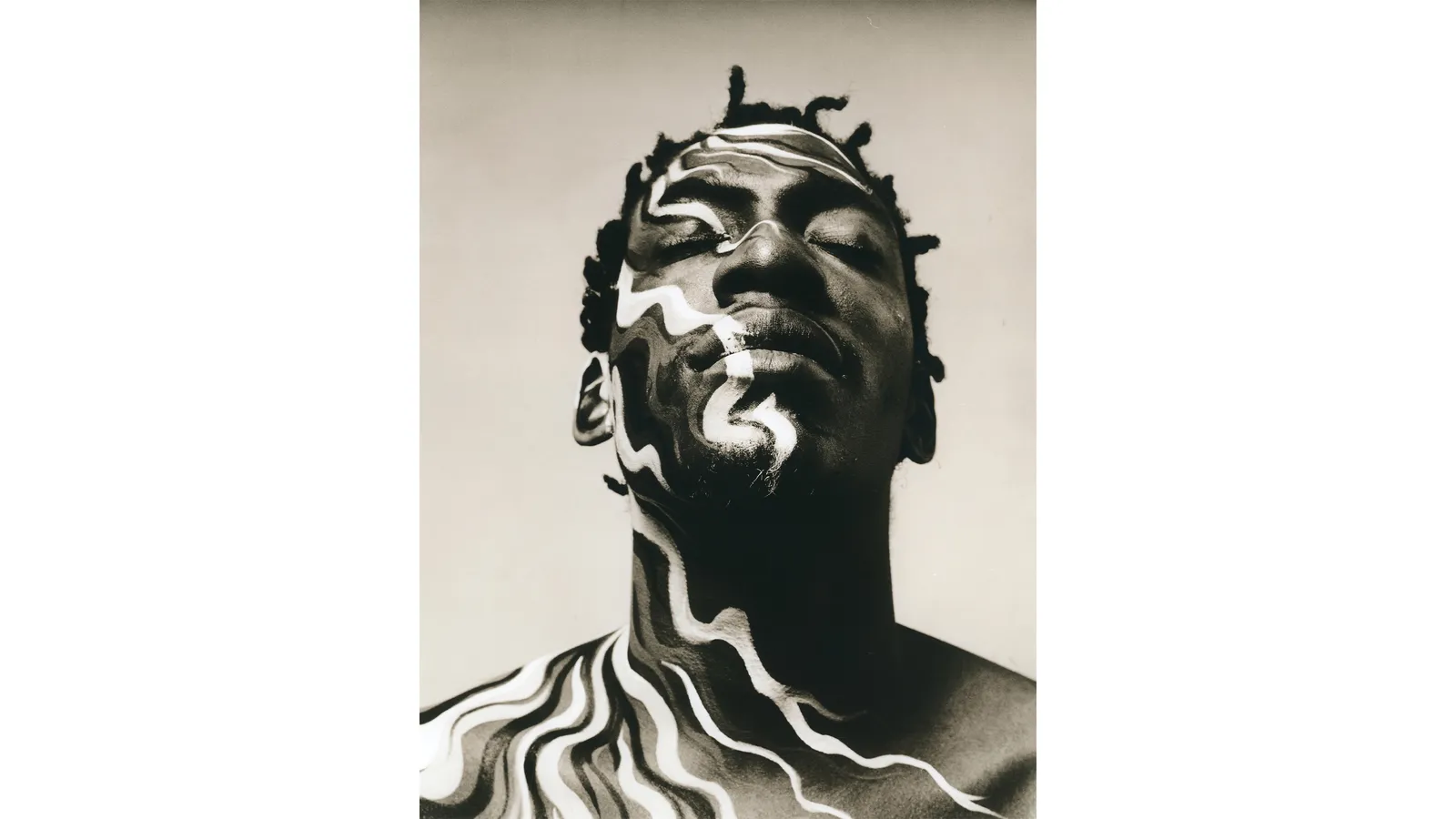 A bust of a man with black and white face paint, showcasing intricate details of the human face, including the jawline. Behind the bust is a building that complements the artistic theme.