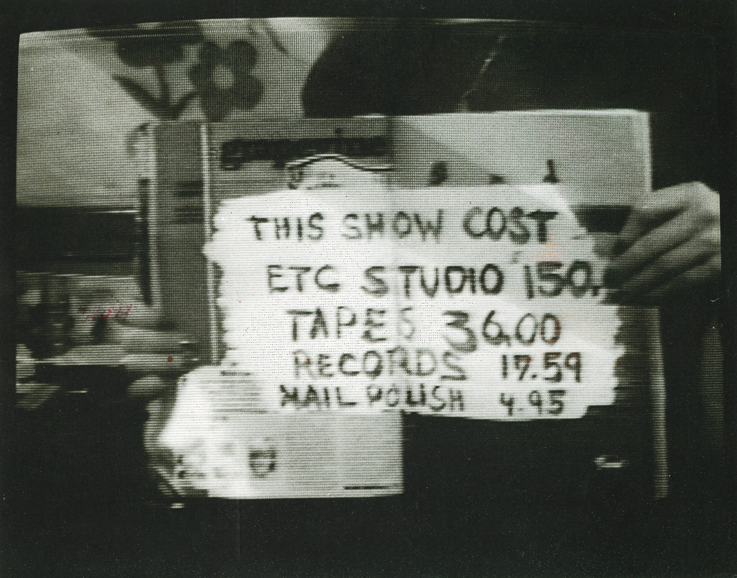 A person holding a sign with handwritten text that lists various prices: "THIS SHOW COST ETC STUDIO 150. TAPES 36.00 RECORDS 19.59 NAIL POLISH 4.95." The image is in black and white, showcasing the sign and the individual on a street.