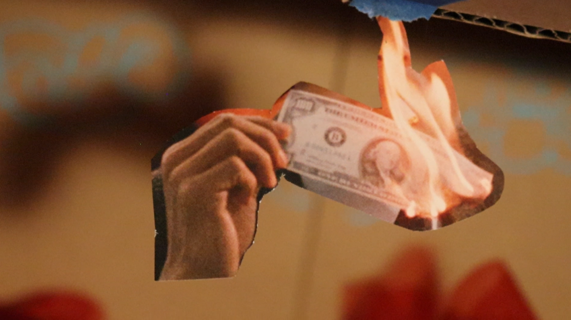 A hand holding a burning piece of paper money indoors.