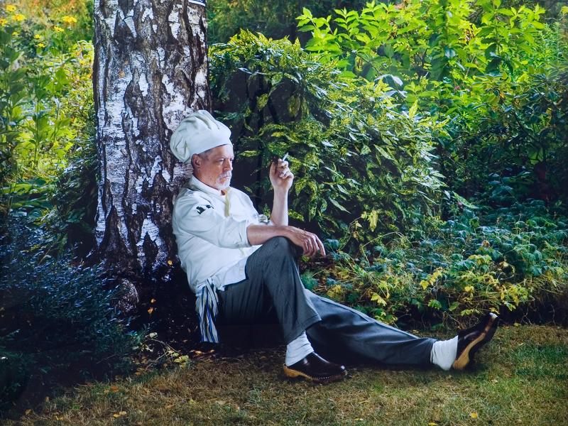 A man sitting outdoors next to a tree, wearing clothing and footwear, on grass.