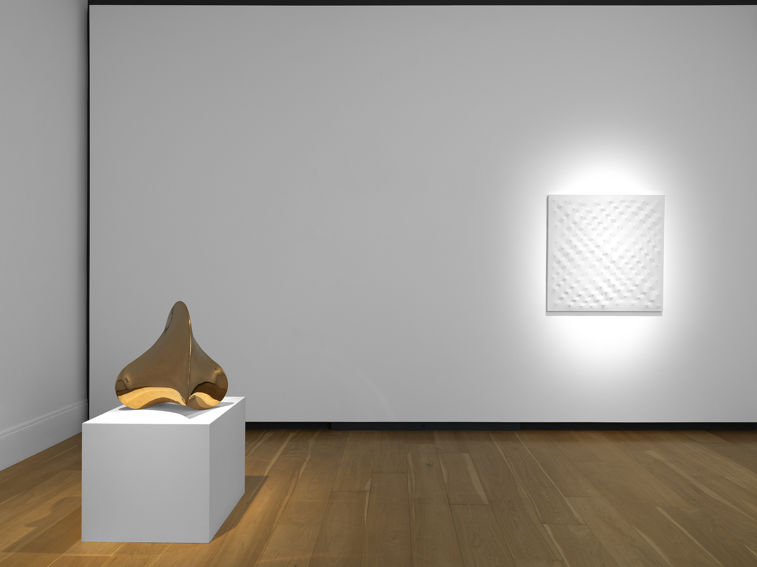 A polished bronze sculpture with curved forms sits on a white pedestal, beside a textured white artwork on a gallery wall.