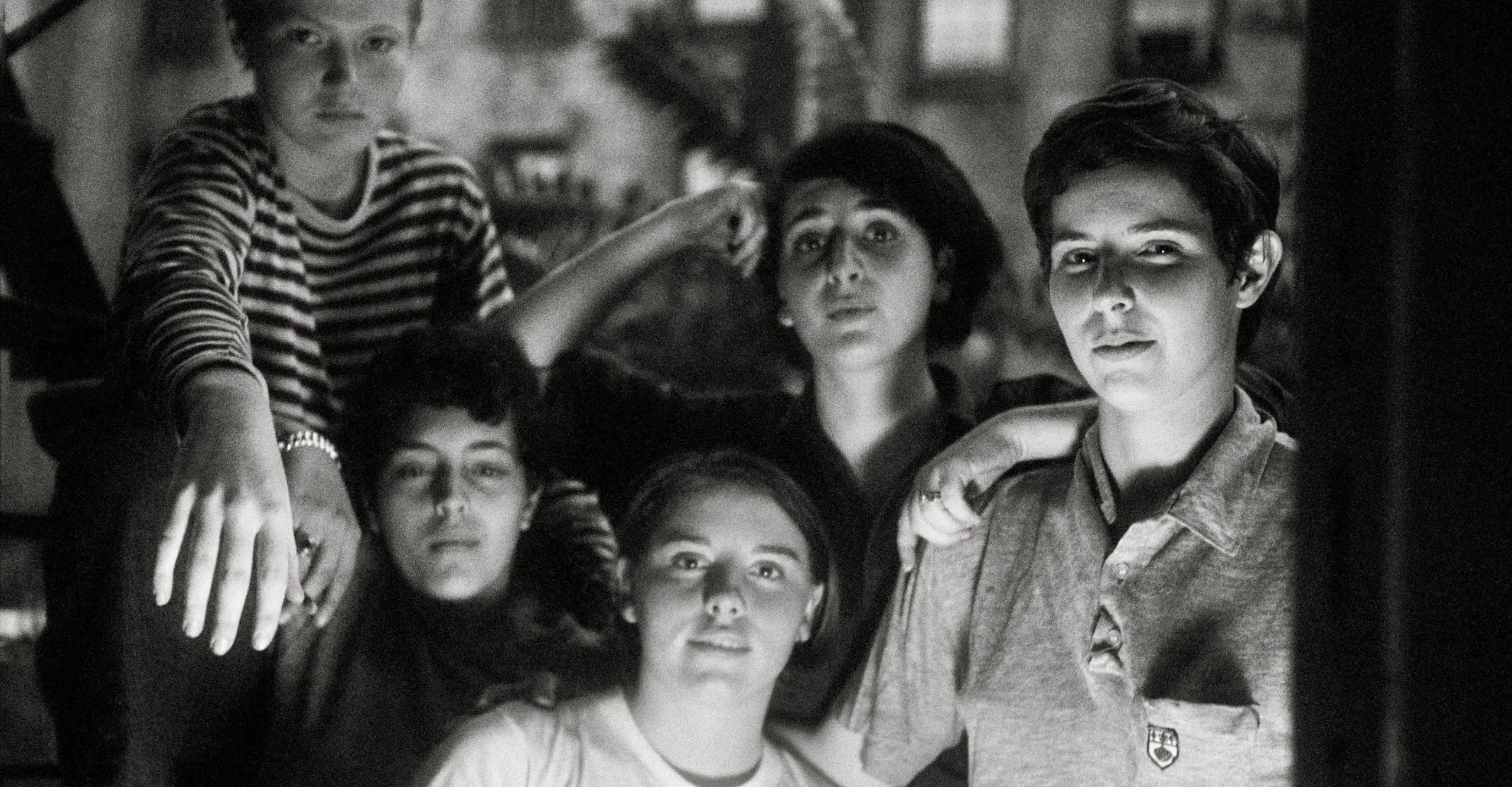 Five individuals are standing together in a dimly lit setting, with two of them positioned in the front and three behind. The group has a serious expression, and their clothing styles vary. The background appears to be somewhat blurred, focusing attention on the group.