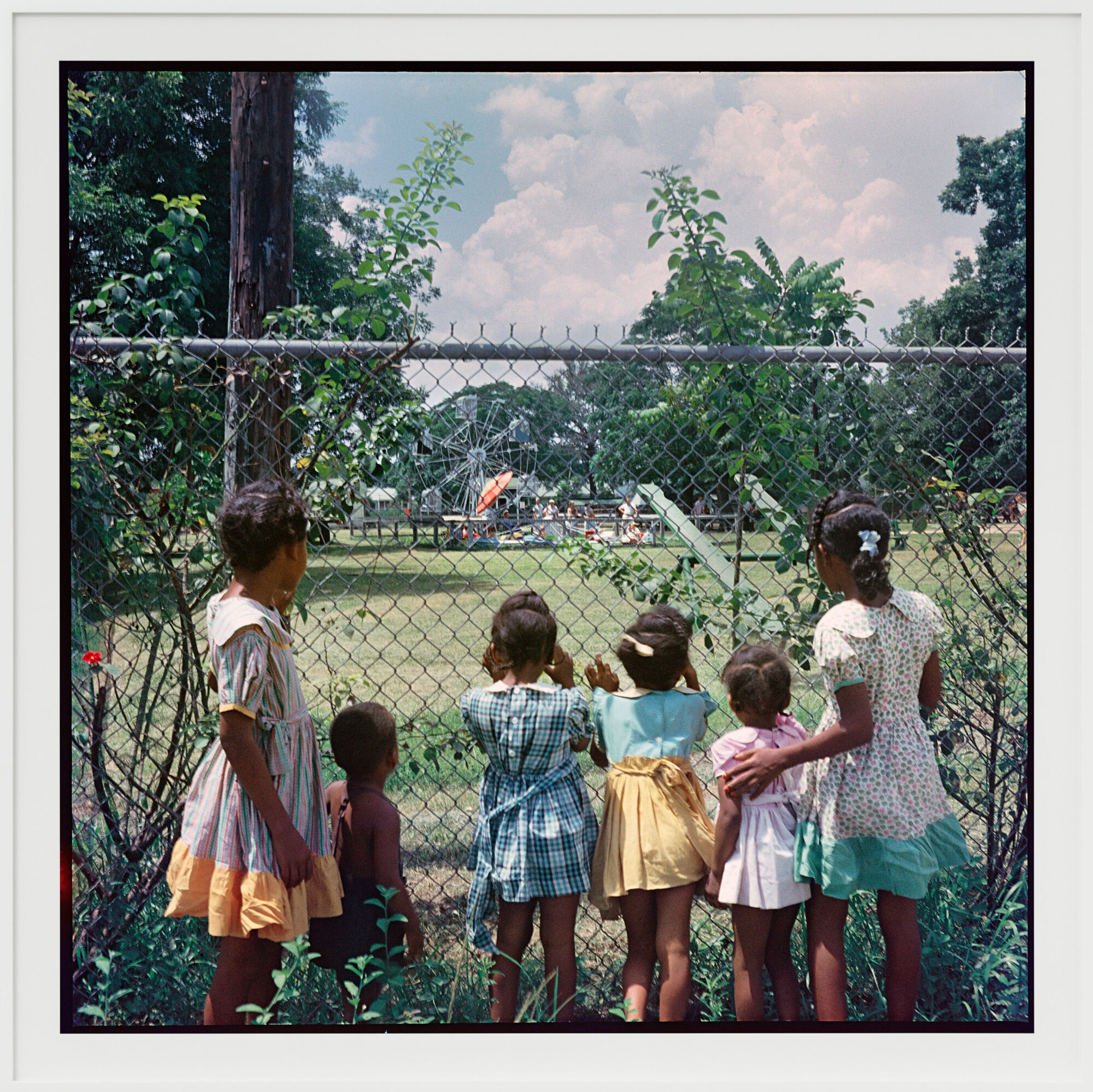 A group of children standing by a fence, looking out towards a playground or park area in the distance, surrounded by trees and bright sunlight.