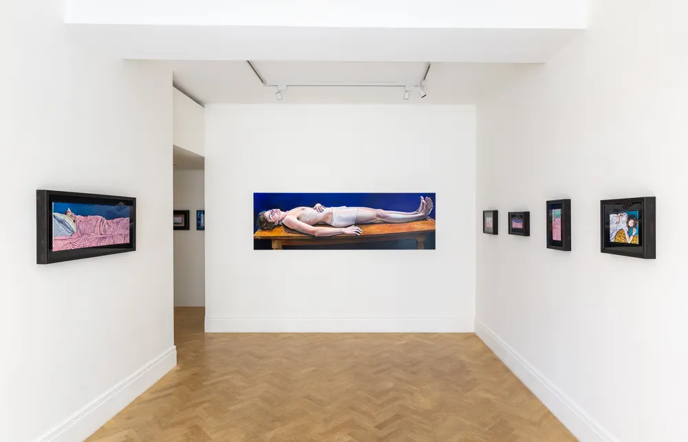 A gallery interior featuring a long horizontal artwork of a figure lying down on a table, surrounded by smaller framed artworks on the walls. The space is well-lit with white walls and light wooden flooring.