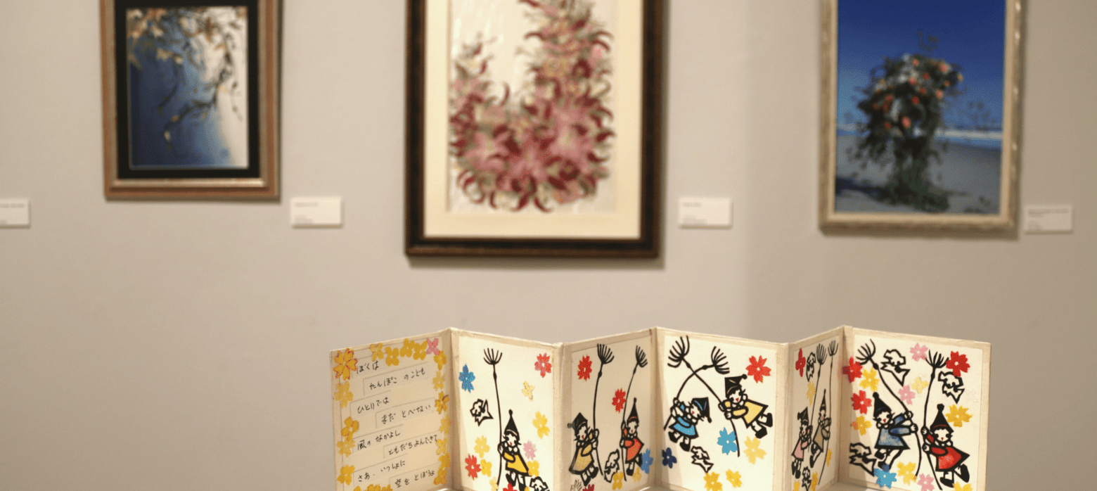A display of three artworks on a gallery wall, with an open book featuring illustrations and handwritten text in the foreground.