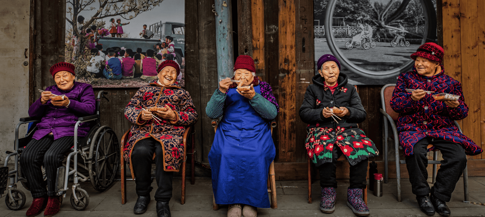 Five elderly women sitting together in traditional clothing, holding bowls of food, with a colorful backdrop featuring a black-and-white photograph.