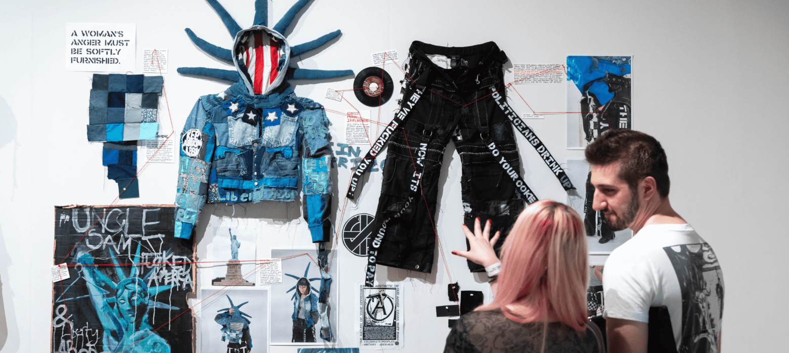 A gallery exhibition showcasing a blue and black outfit resembling the Statue of Liberty, alongside various artistic elements including posters and graffiti. Two viewers are examining the artwork.
