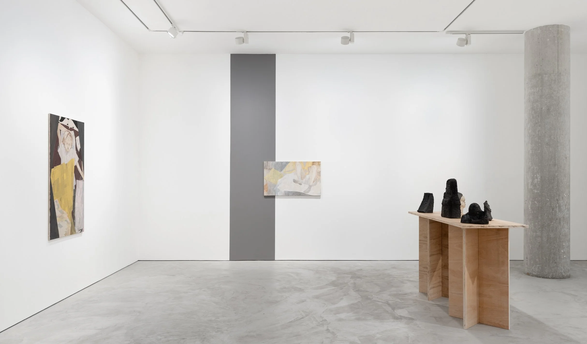 An art gallery interior featuring abstract paintings on white walls, a gray vertical strip, and a wooden table displaying sculptural objects.