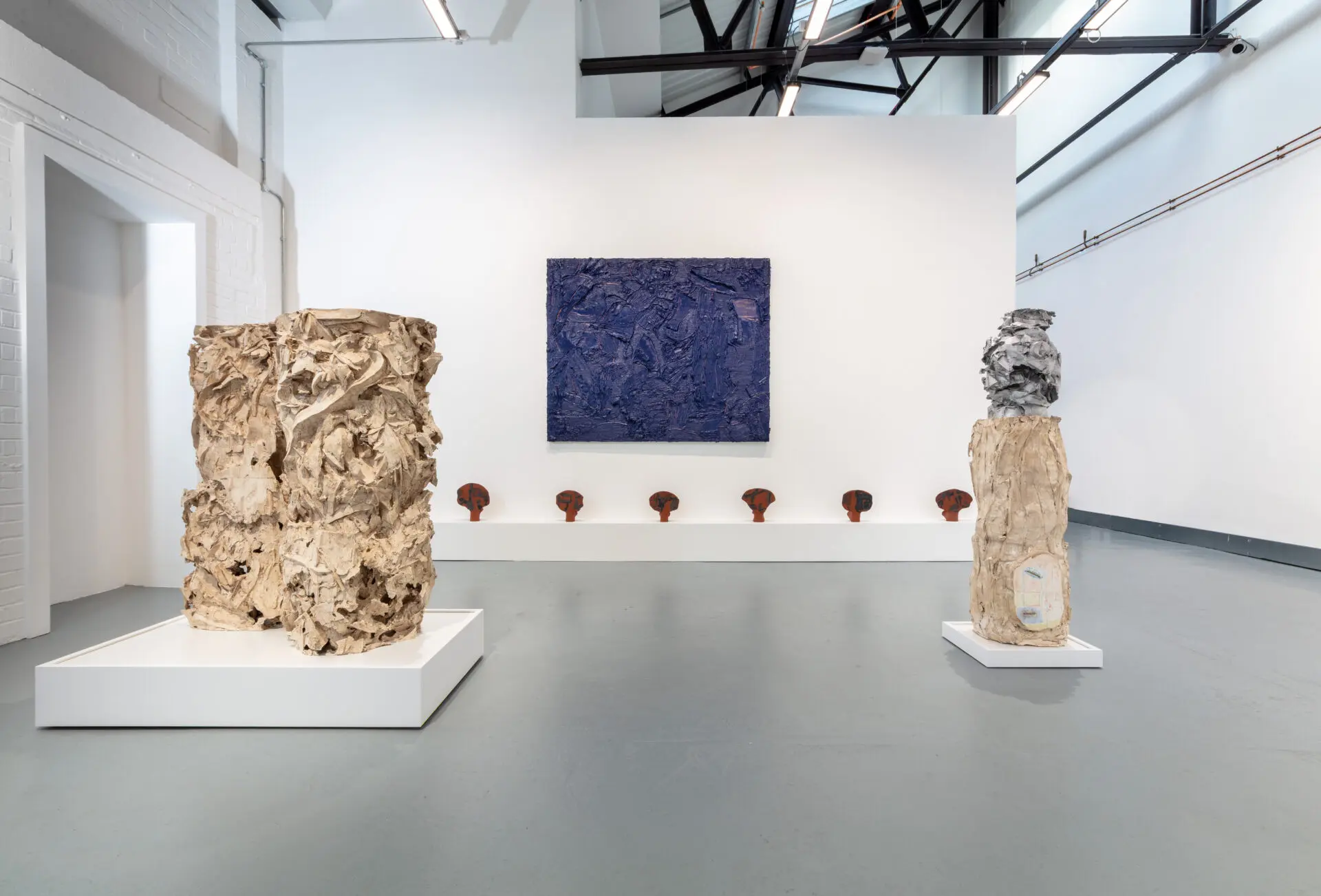 A contemporary art gallery featuring abstract sculptures on pedestals, with a large textured blue painting on the wall and smaller artworks below it.