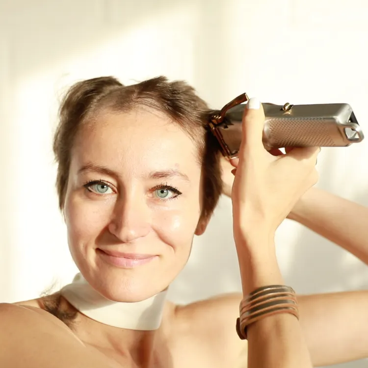 A woman with long hair is holding a hair clipper, smiling as she prepares to style her hair. She has bright blue eyes and is wearing a choker necklace.
