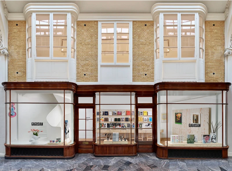 A view of a gallery exterior with large glass windows displaying books and art pieces, featuring wooden accents and high ceilings.