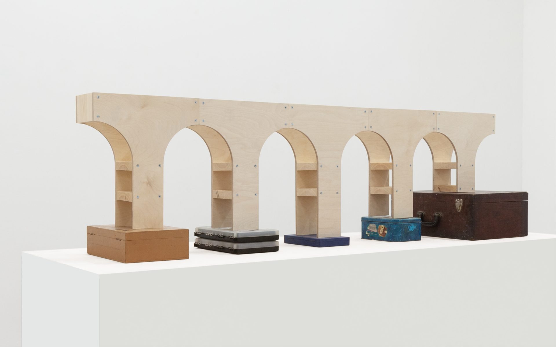 A minimalist sculpture featuring a series of arch-shaped wooden structures mounted on a white pedestal, with several boxes of varying sizes and colors placed underneath the arches.
