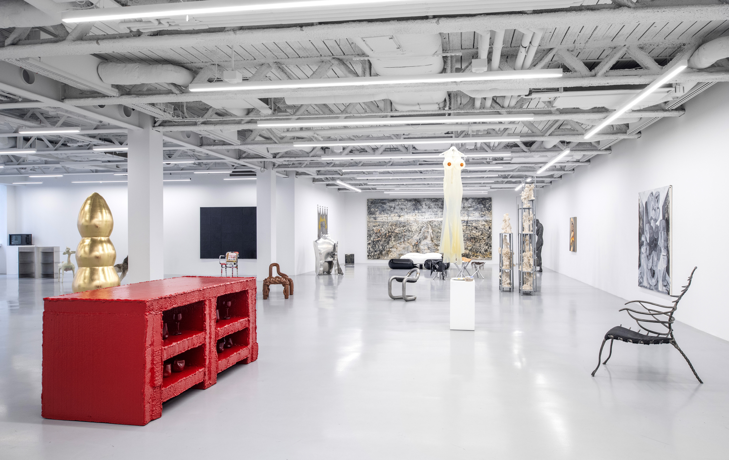 A spacious art gallery with white walls and a polished floor. Various artworks are displayed, including sculptures and paintings. There is a large red sculptural piece made of blocks, and several other distinct objects scattered throughout the room.