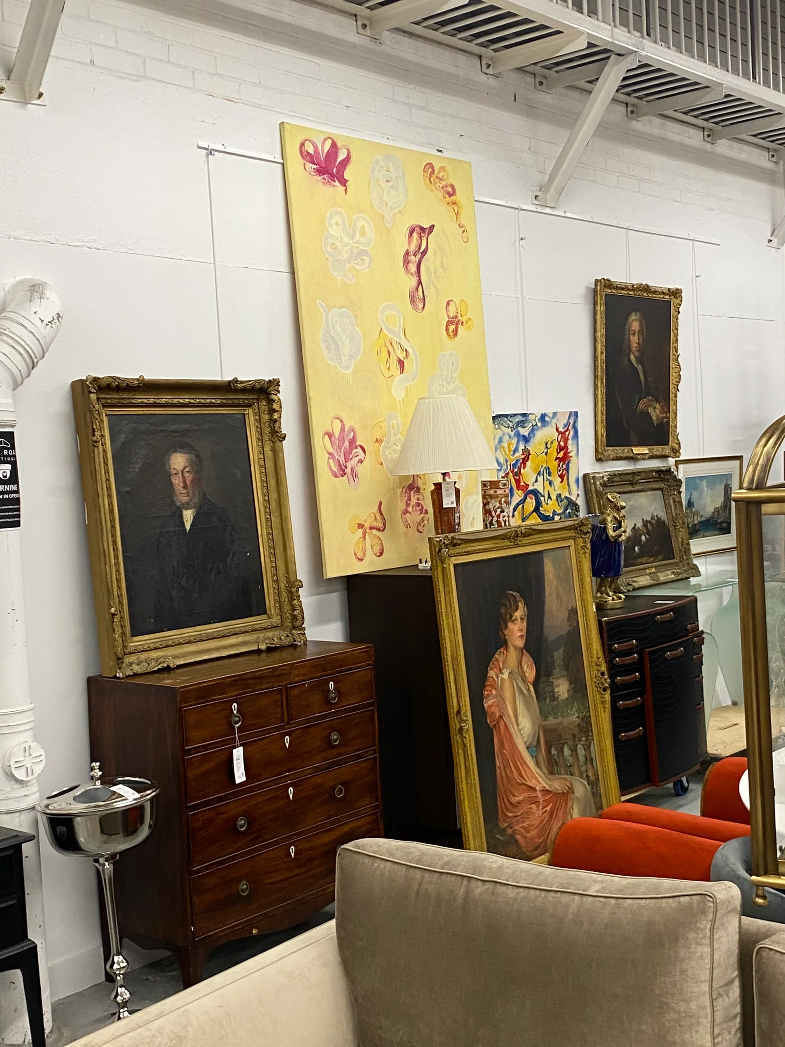 Various pieces of art and furniture are displayed in a gallery-like setting, including ornate framed portraits, a colorful abstract painting, and vintage furniture items like a chest of drawers.