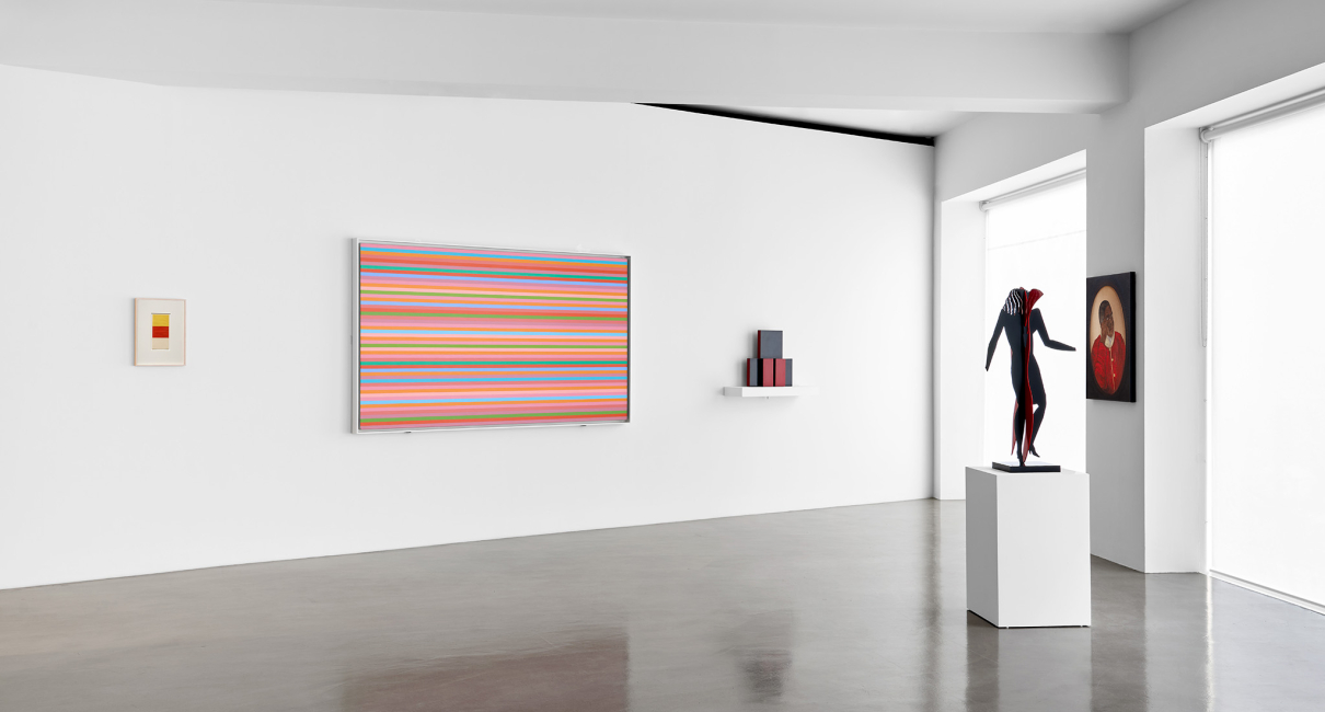 A modern art gallery featuring a variety of artworks, including a colorful striped painting, a red and black sculpture of a figure on a pedestal, and framed pieces on the walls. The space has large windows allowing natural light to illuminate the room.