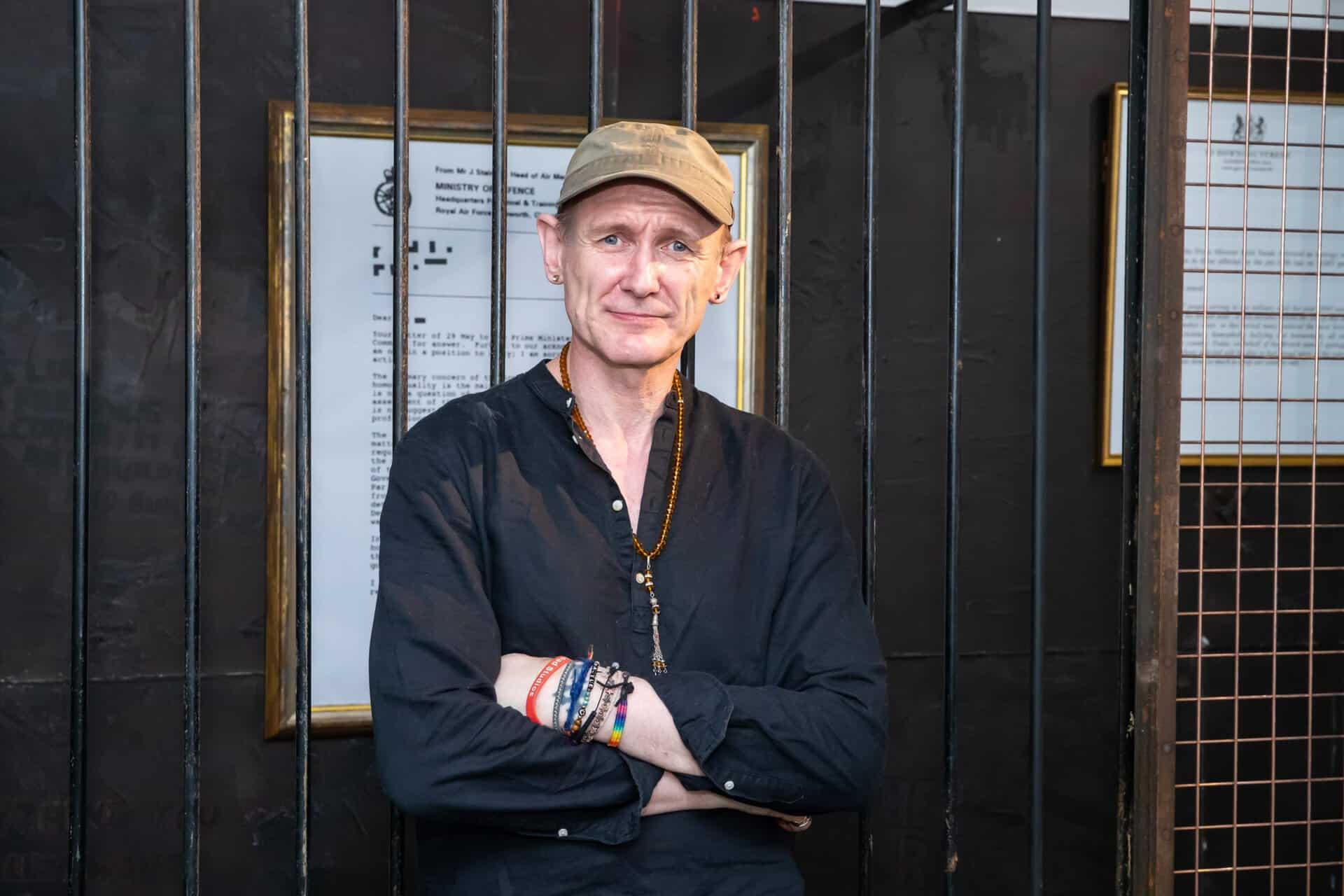 A man wearing a cap and black shirt stands with folded arms behind black metal bars, with framed documents on the wall behind him.