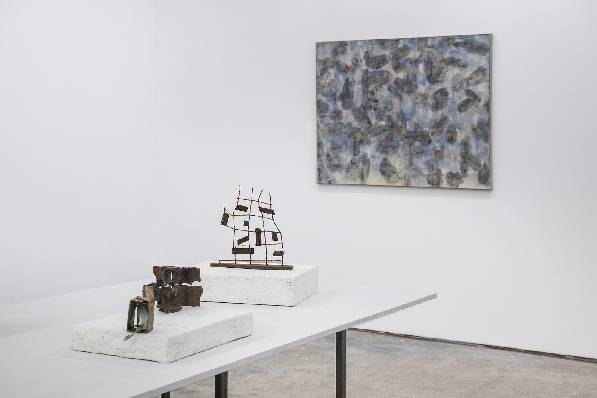Minimalist gallery with a blue-gray abstract painting, two small sculptures, and a model sailing ship on white pedestals; serene mood.