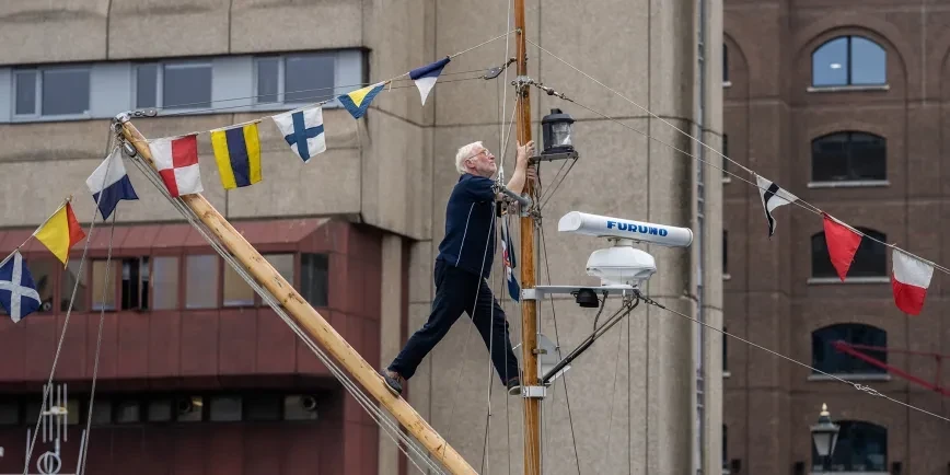 Sailor climbs a wooden mast beside a building, surrounded by colorful nautical flags, rigging, and radar equipment in a maritime display.