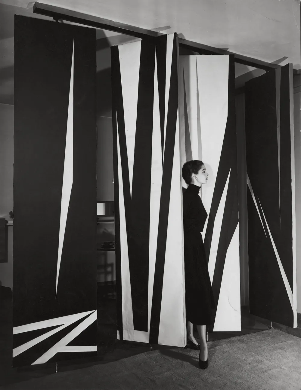 Black-and-white avant-garde fashion scene: a woman in a dark dress stands among tall geometric panels and sharp abstract shadows, creating drama.